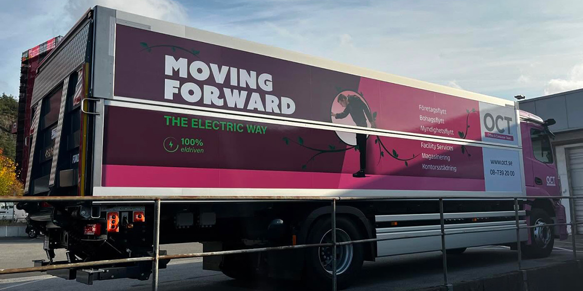 Rosa OCT-lastbil med texten ’Moving Forward – The Electric Way’ parkerad vid en byggnad.